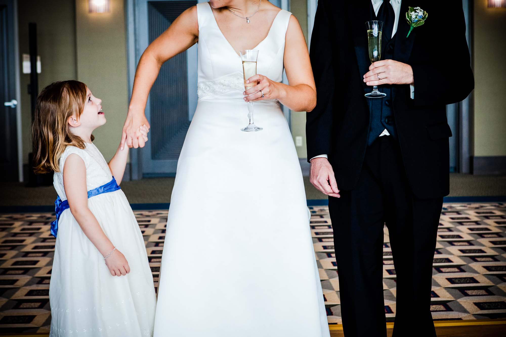 little girl in flower girl dress smiles up at the bride and groom and holds the bride's hand as they are holding champagne glasses in a reception room