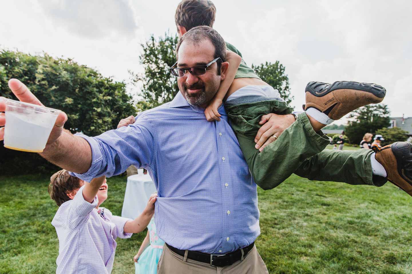 man holds kid over his shoulder while another one pulls on his shirt, sunglasses disheveled and holding a drink out of the picture