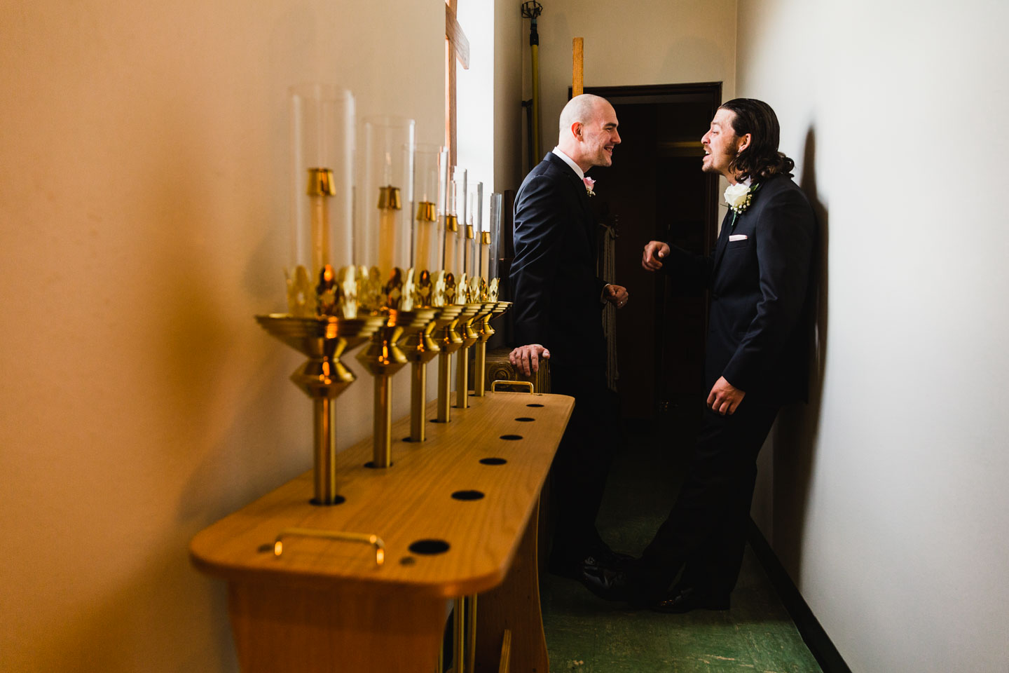 candid moment between a groom and his groomsman, in the back of the church, before the ceremony, surrounded by candles in a narrow hallway